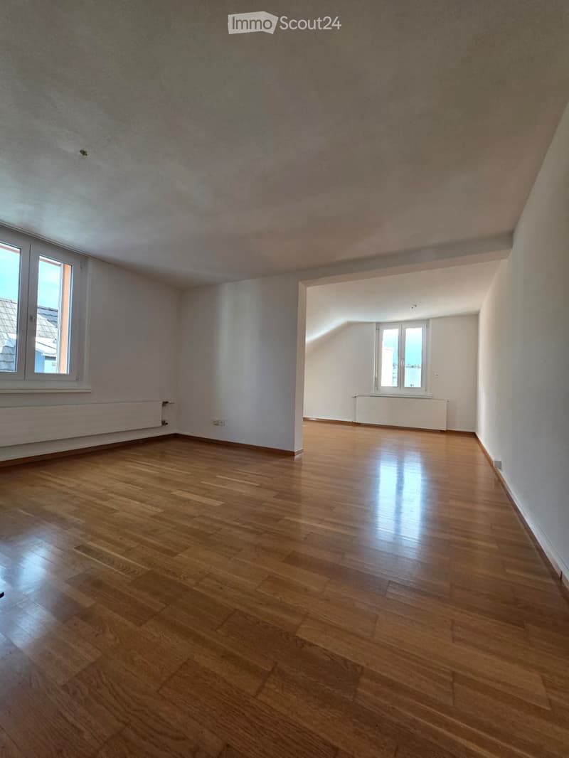 Empty room with wooden flooring, white walls, and large windows allowing natural light. Radiators are visible under the windows.