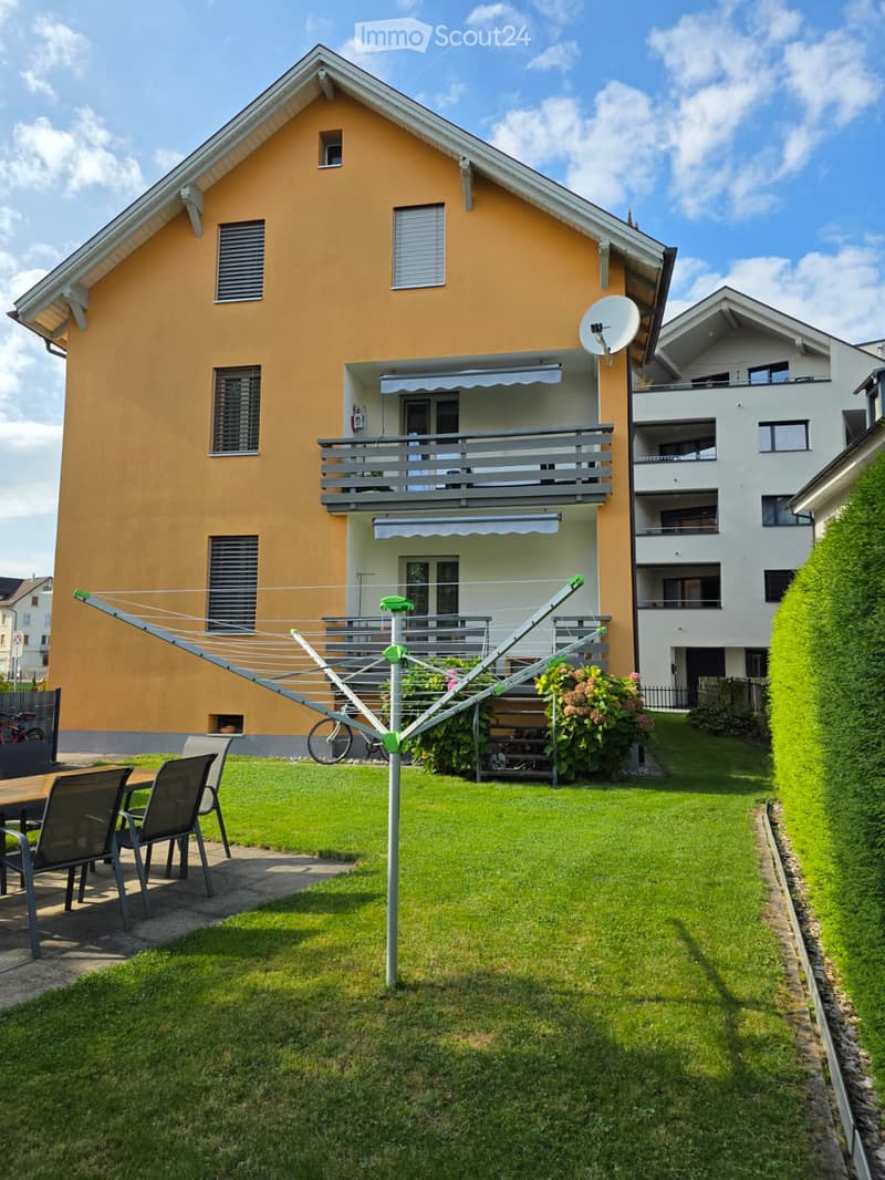 A well-maintained garden area in front of a two-story house with a balcony, featuring a clothesline, outdoor furniture, and a bicycle.