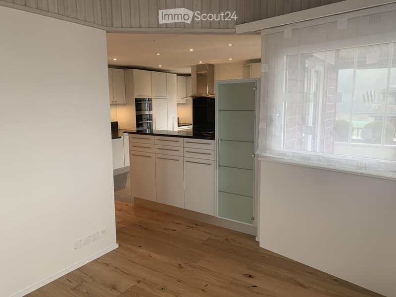 Modern kitchen with white cabinets, wooden flooring, and a large window with sheer curtains. Features include an oven, range hood, and drawer units.