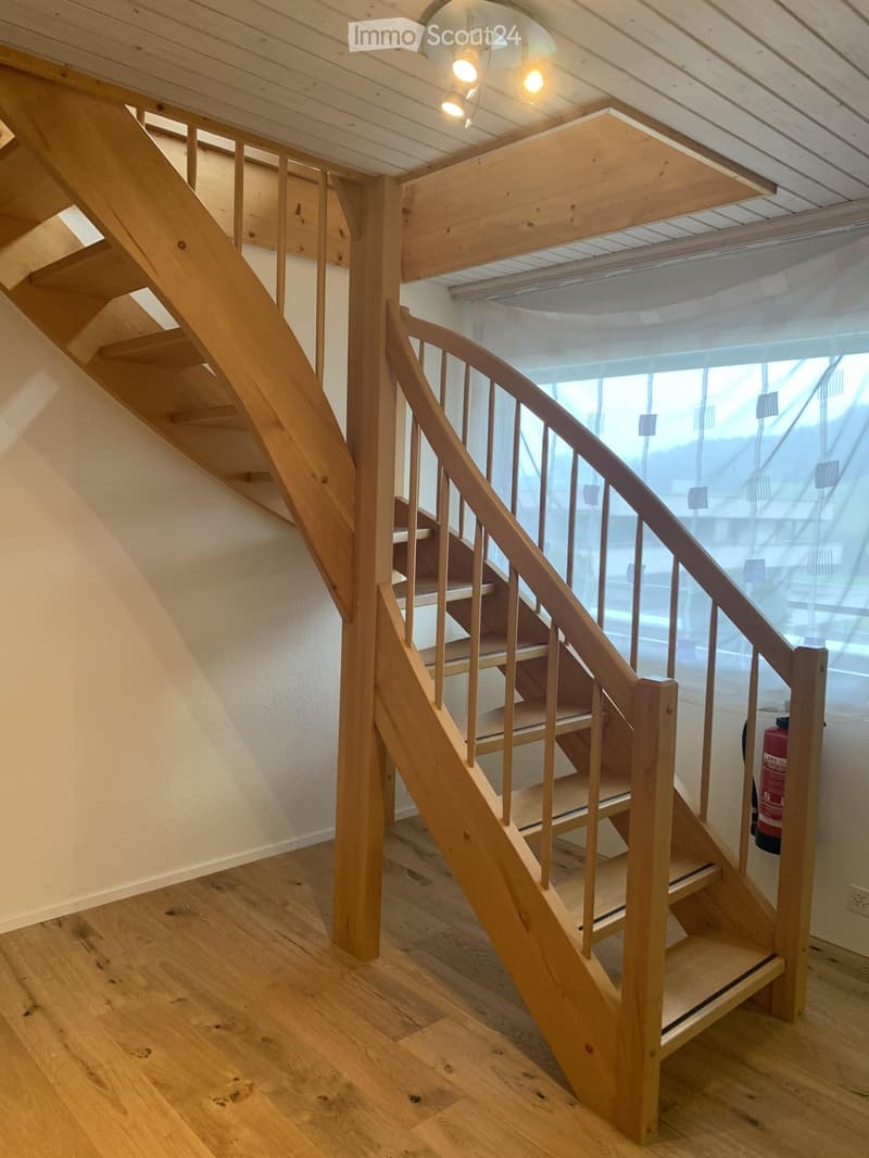 Wooden staircase with a curved handrail and open wooden balustrade, leading to an upper level. The staircase is illuminated by ceiling lights, and the walls are painted white. A red fire extinguisher is visible on the right side.