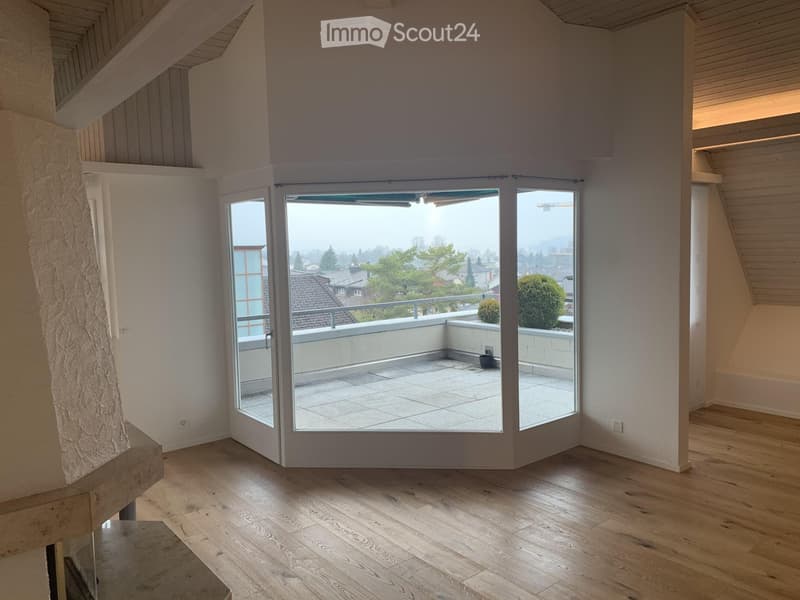 Bright, empty room with wooden flooring, large sliding glass doors leading to a tiled balcony, and a view of the city and surrounding greenery.
