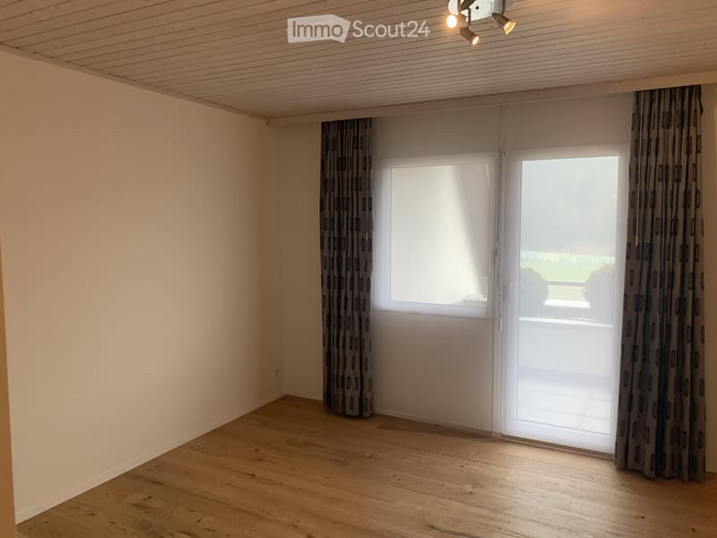 Empty room with wooden flooring, white walls, large window with curtains, and a sliding glass door leading outside.
