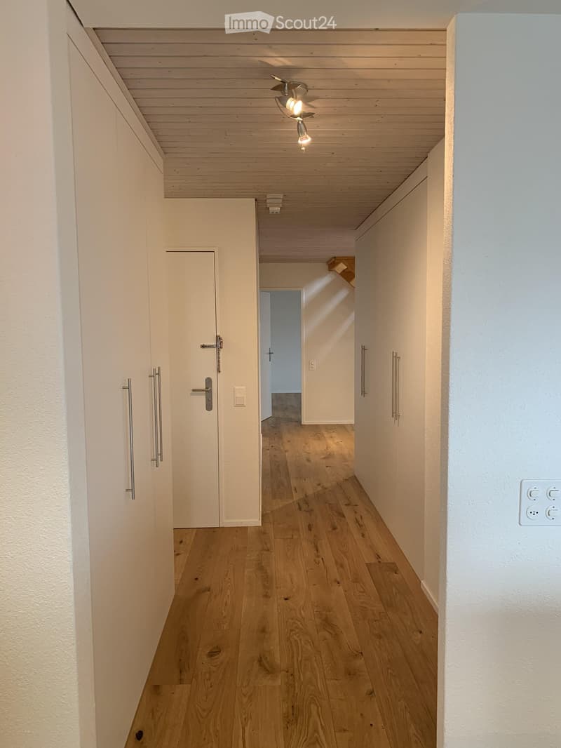 Clean, modern hallway with light wooden flooring, white walls, and a ceiling light fixture. Features a closed door on the left and an open doorway leading to another room at the end.