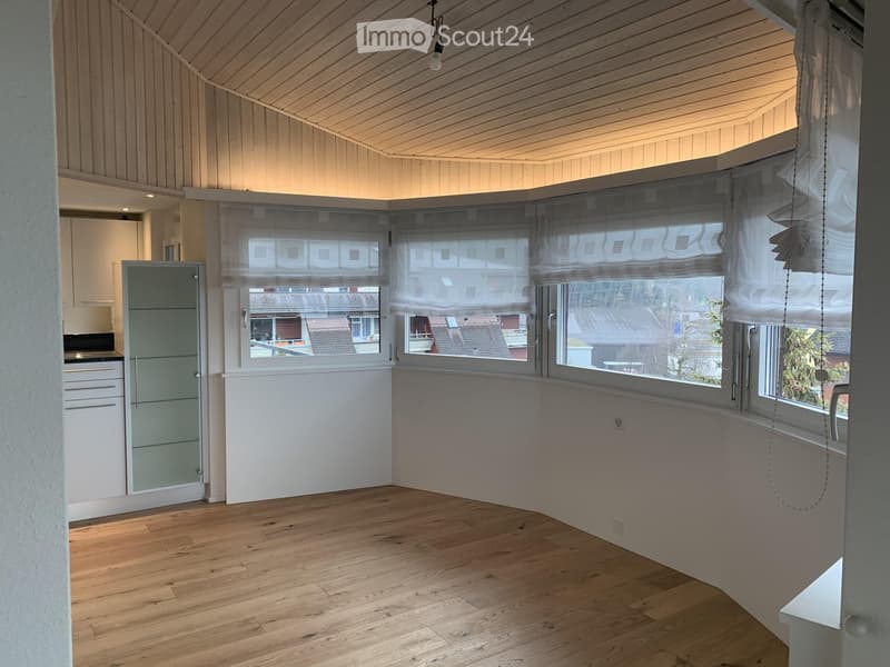 Bright, empty room with wooden flooring, large windows with blinds, and a view of neighboring buildings. The room features white walls and a simple, modern design.