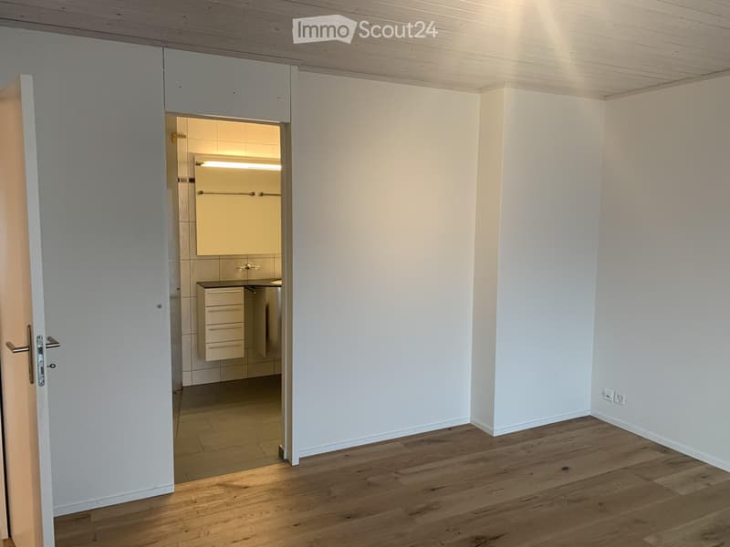 Empty room with wooden flooring and white walls, featuring an open doorway leading to a small, modern bathroom with white cabinets and a sink.