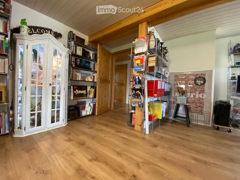 A cozy, well-lit room with wooden flooring and exposed wooden beams. Features include a white cabinet with glass doors, a bookshelf filled with books and decorative items, a small stool, and a cart with storage boxes.