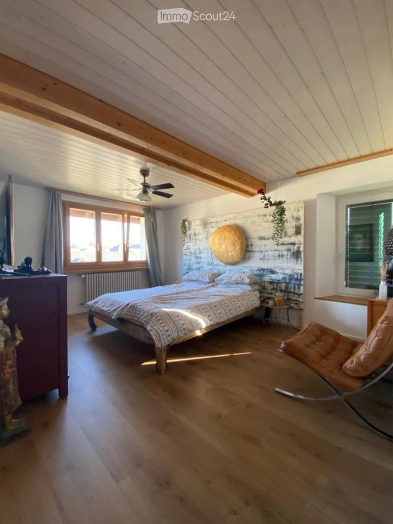 Cozy bedroom with wooden floors, exposed wooden beams on the ceiling, large windows with grey curtains, a bed with colorful bedding, a nightstand, a modern chair, and a decorative wall piece.