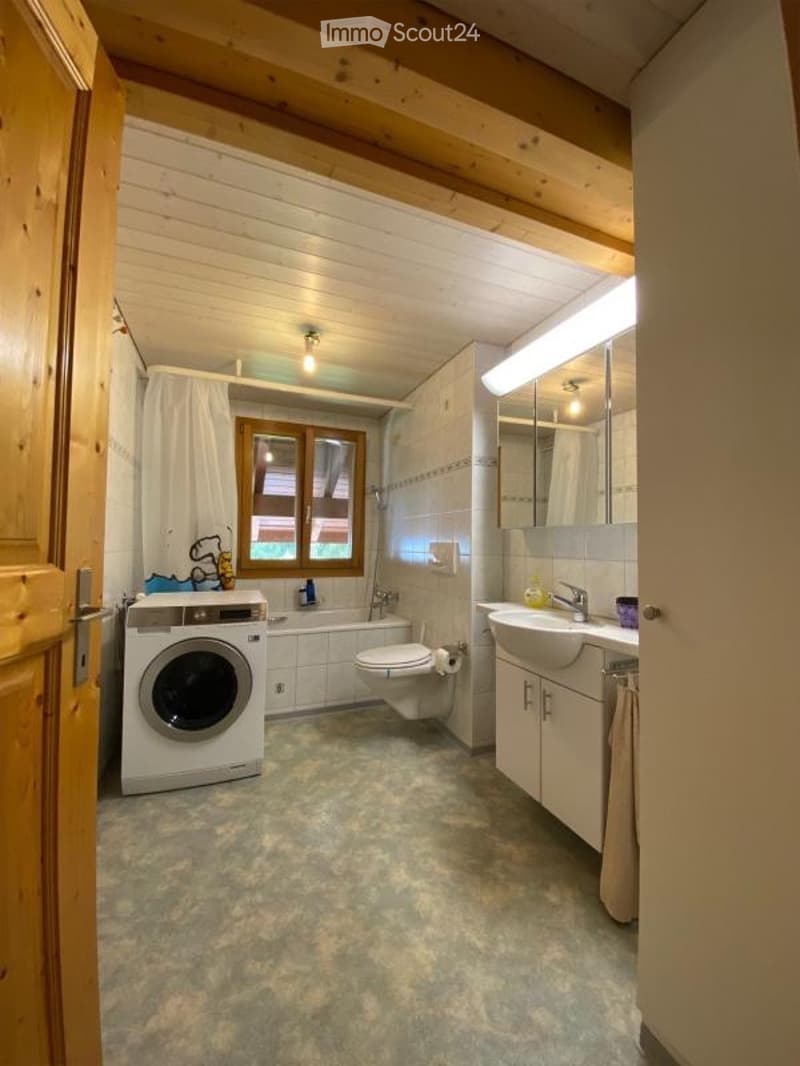 Bathroom with wooden ceiling beams, a window, a sink with a mirror, a toilet, a bathtub, and a washing machine. The room features a light fixture and a curtain on the window.