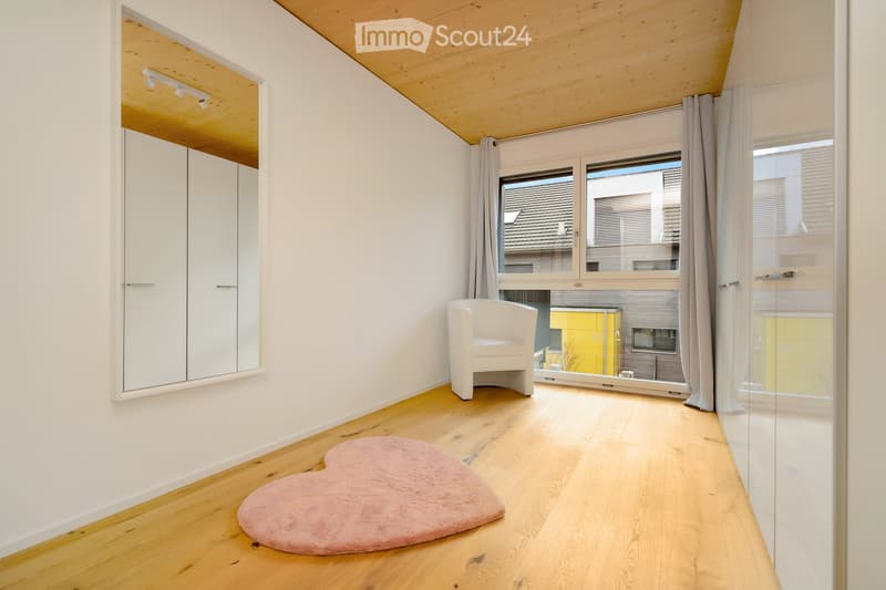 Empty room with wooden floor, white walls, a mirror, and a heart-shaped rug. There is a white chair and a glass window with curtains.