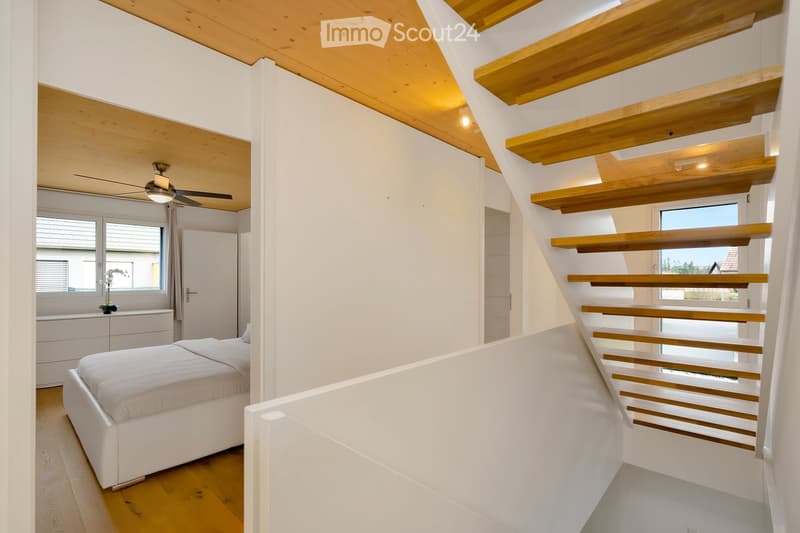 Modern bedroom with white bed, wooden stairs, and white walls. Features a ceiling fan and a window with a view.