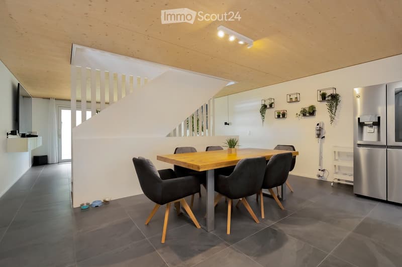 Modern dining area with a wooden table, black chairs, and a potted plant. The room has a white wall, a ceiling with lights, and a refrigerator on the right side.