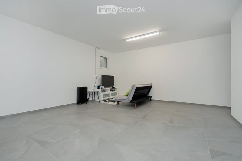 An empty room with white walls, gray tiled floor, a white entertainment center, a black television, a black radiator, a gray chaise lounge with a green pillow, and a small table.