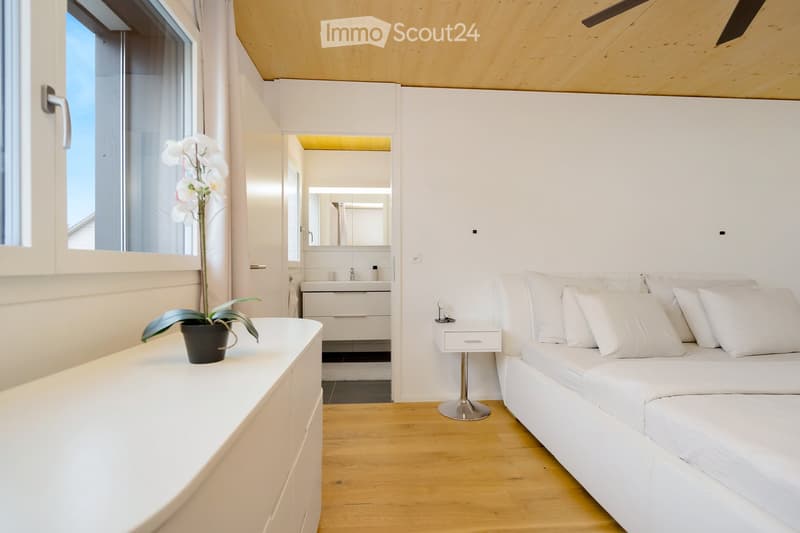 Minimalist bedroom with white walls, wooden floor, and a bed with white bedding. A nightstand with a plant and a cabinet with a sink and mirror are visible. The ceiling has a wooden design and a ceiling fan.