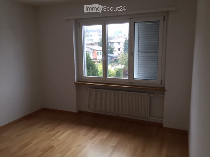Empty room with wooden flooring, large window offering a view of the neighborhood, radiator below the window, and white walls.