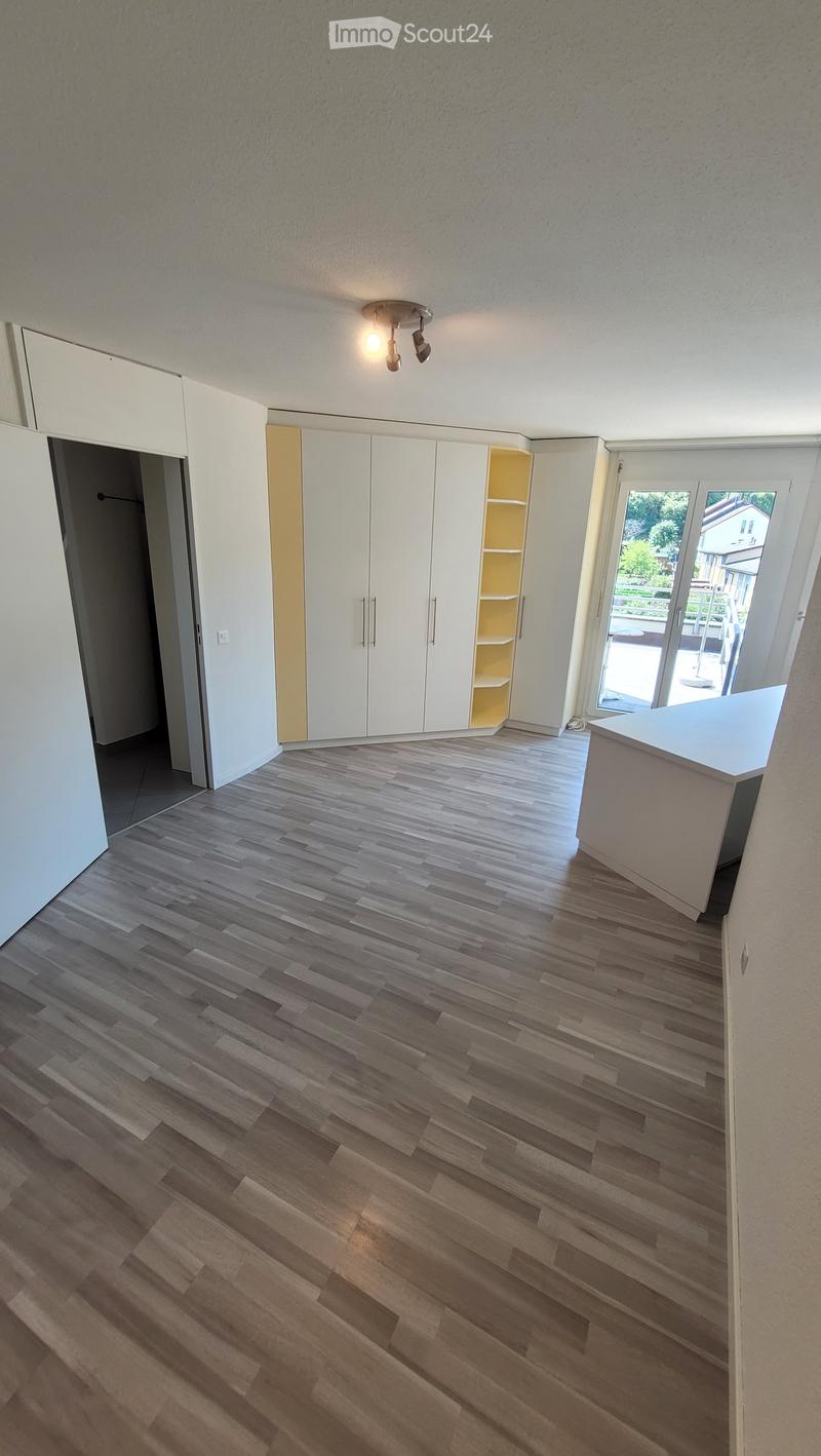 Empty room with wooden flooring, white walls, built-in white cabinets, and a large window with a view of the garden.