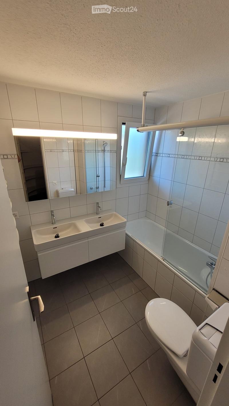 Modern bathroom with white tiles, double sink, toilet, bathtub with glass enclosure, and large mirror with built-in lighting.
