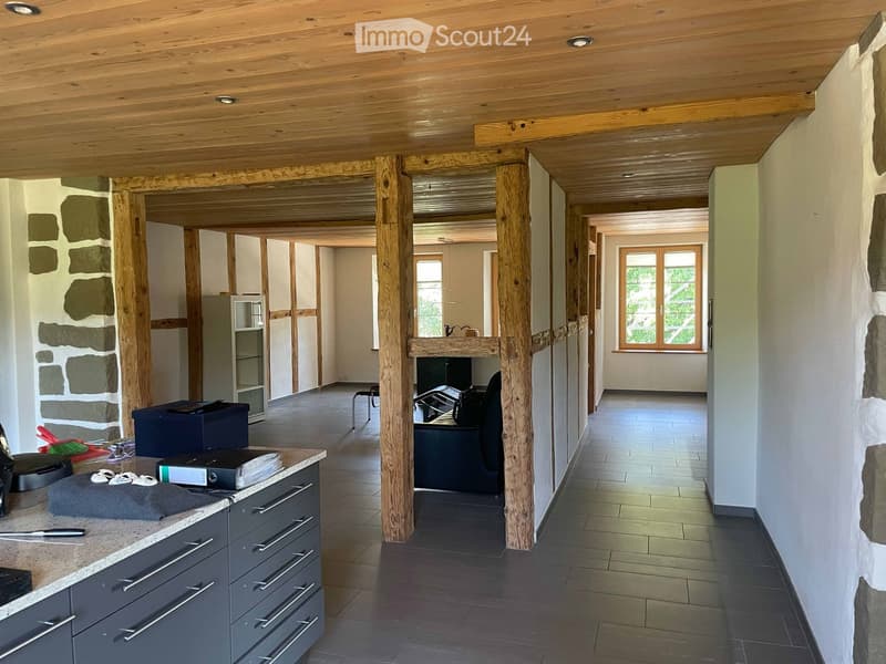 A modern, open-plan office space with wooden beams, a desk, chairs, and a cabinet. The room features a polished tile floor and large windows allowing natural light.