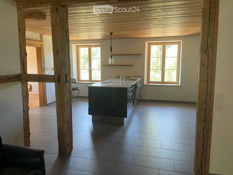Modern kitchen with a central island, sink, and pendant light. Large windows provide natural light, and the space features a wooden ceiling and tiled floor.
