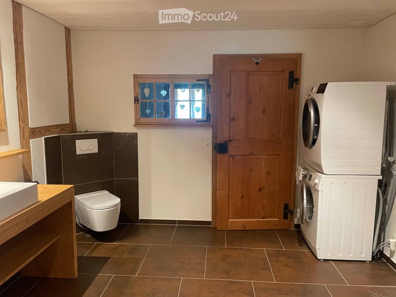 Bathroom with a toilet, sink, and stacked washing machine and dryer. Features a wooden door, window with decorative glass, and tiled floor.