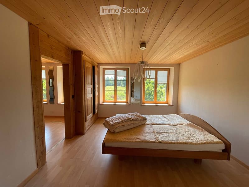 Bright bedroom with wooden floors, slanted wooden ceiling, large windows, and a simple wooden bed with white bedding.