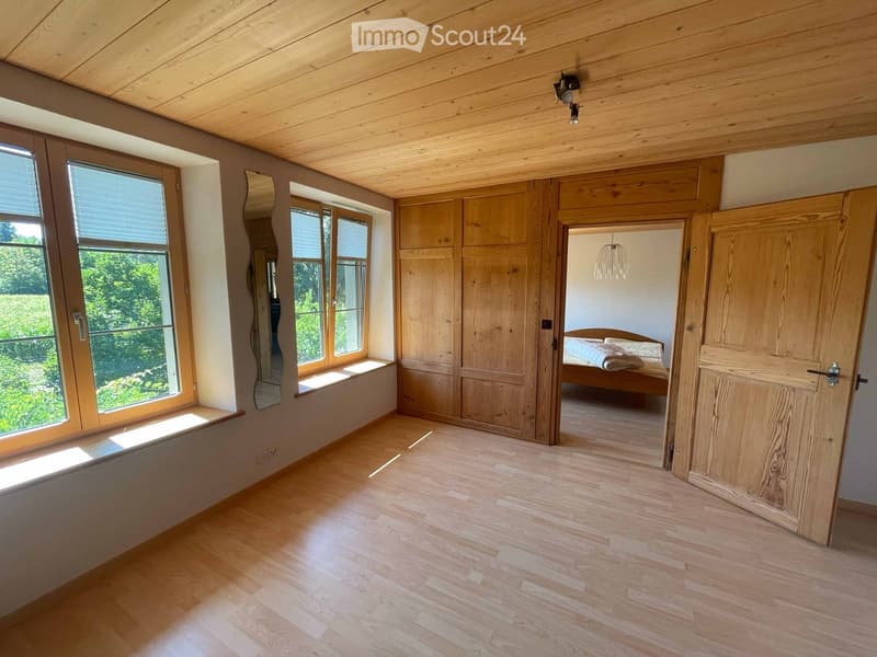 Empty room with wooden flooring, large windows, built-in wooden cabinetry, and a doorway leading to a bedroom.