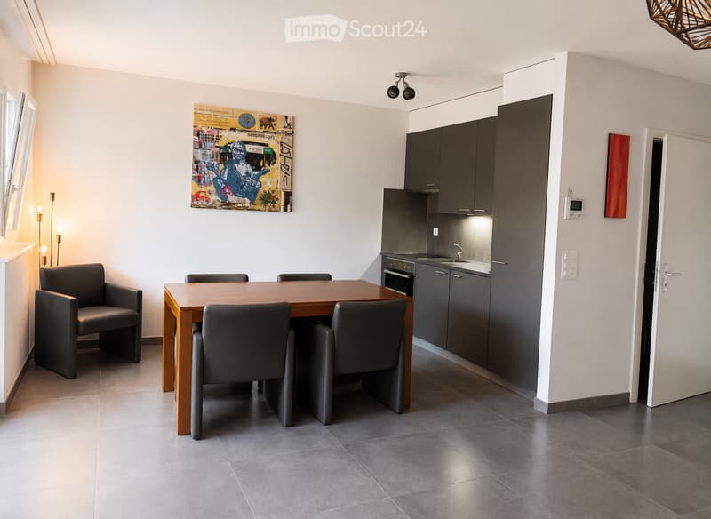 Modern open-plan living and dining area with a wooden dining table, four grey chairs, grey cabinets, a sink, and a small oven. The space features a large abstract painting on the wall and a window with blinds.