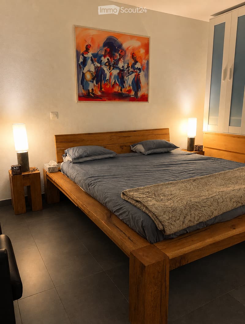 A modern bedroom with a wooden bed, nightstands, lamps, a large mirror, and a wardrobe. The bed is neatly made with pillows and a blanket, and a colorful abstract painting hangs on the wall.