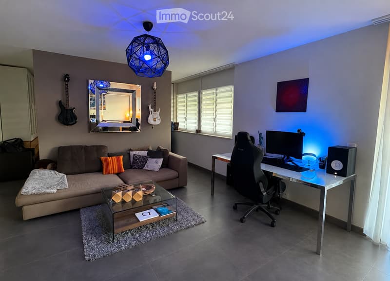 Modern living room with a sofa, coffee table, desk, computer, and guitars on the wall. Features large windows with blinds and a stylish geometric pendant light.