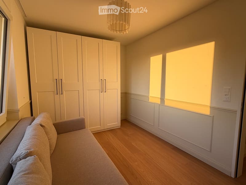 A modern living room with a grey sofa, a large white wardrobe, and a hanging chandelier. The room features wooden flooring and large windows with blinds.