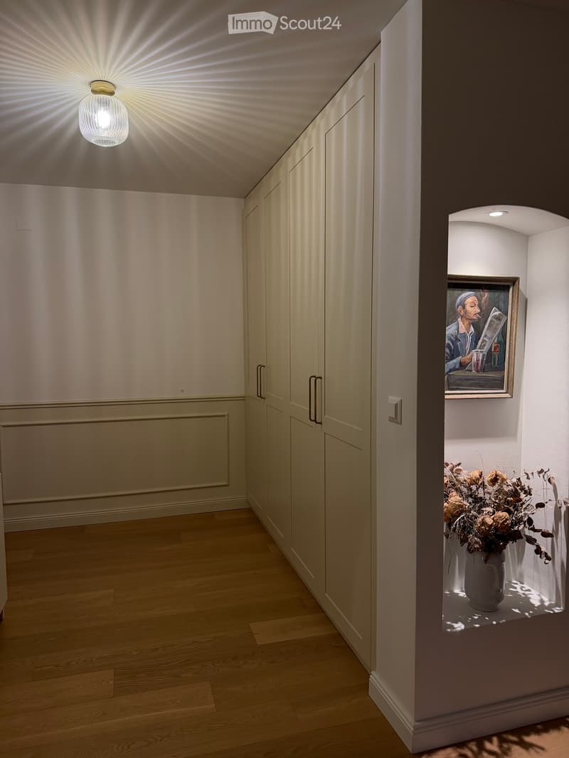 Modern hallway with light wooden flooring, white walls, built-in closet doors, and a small niche with a framed painting and a vase of dried flowers.