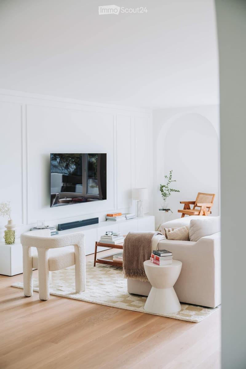 Bright living room with white walls, wooden flooring, and large windows. Features a cream-colored sofa, a modern armchair, a small round stool, a coffee table, a television mounted on the wall, and decorative items including books, a lamp, and a plant.