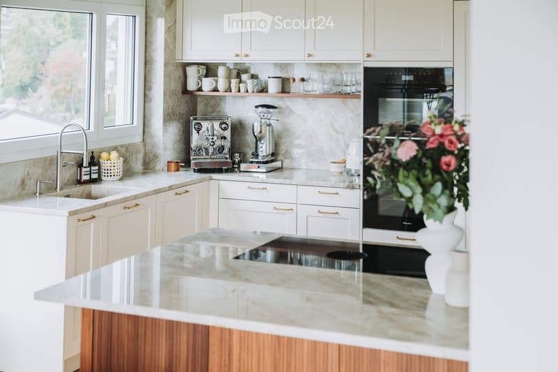 Modern kitchen with white cabinets, marble countertops, stainless steel appliances, and a built-in oven. Features include a sink with a modern faucet, a coffee maker, and decorative items on the counter.