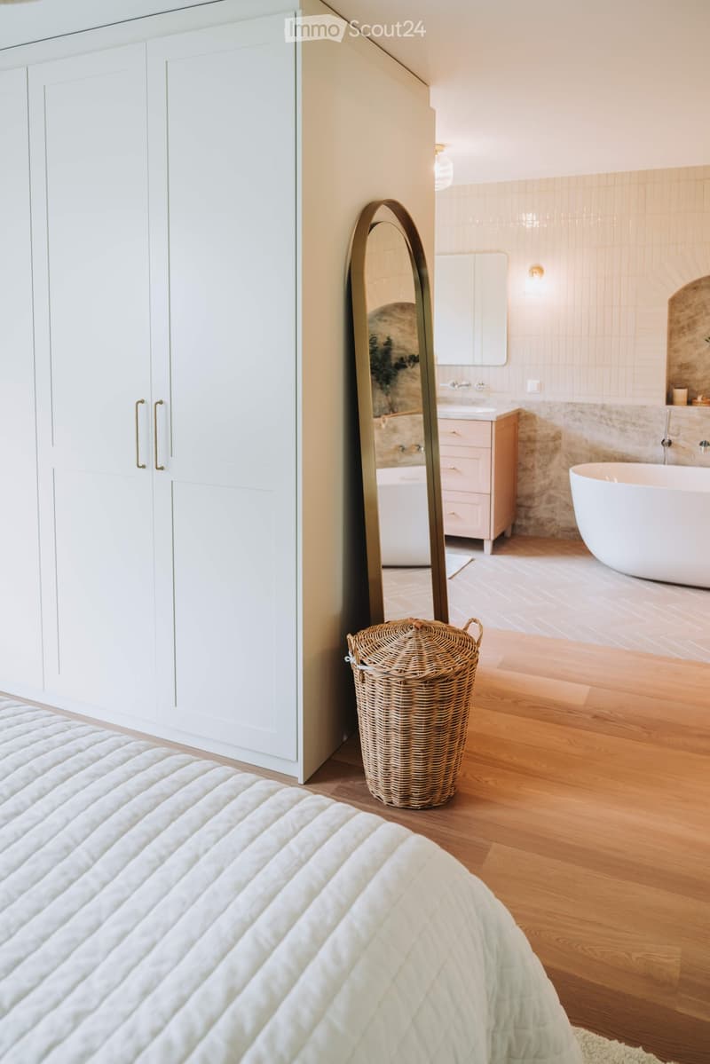 Modern bedroom with a neatly made bed, large white wardrobe, and a stylish mirror. A wicker basket is placed on the wooden floor, and a bathroom area is visible in the background with a bathtub and sink.