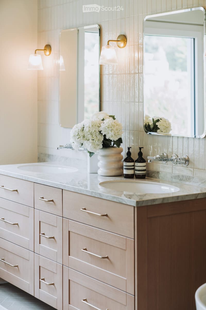 Elegant bathroom with a double sink vanity, marble countertop, light wood cabinets, and a decorative flower arrangement.
