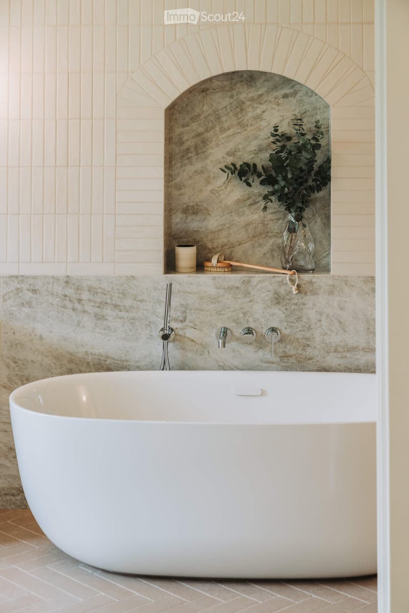 Modern bathroom with a large white bathtub, marble tiles, and a decorative shelf with a vase and brush.