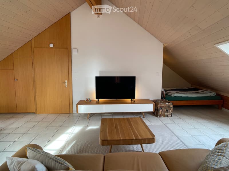 Loft-style living room with a sofa, coffee table, TV stand, television, and a small bed area in the corner.