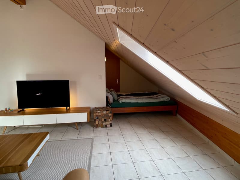 Loft-style room with slanted wooden ceiling, skylight windows, TV on a white console, small bed with green mattress, wooden stool, and a wooden table.