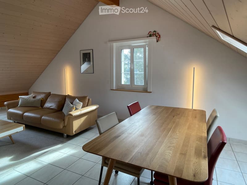 A cozy living room with a slanted wooden ceiling, a large window, a brown leather sofa with decorative pillows, a wooden coffee table, a dining table with chairs, and a floor lamp.