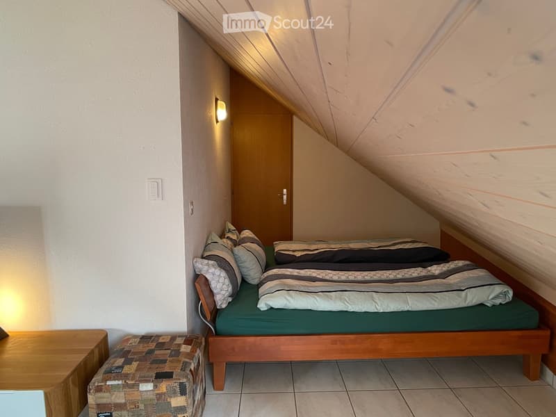 Cozy attic bedroom with a single bed, nightstand, and a small patterned stool. The room features slanted wooden ceilings and warm lighting.