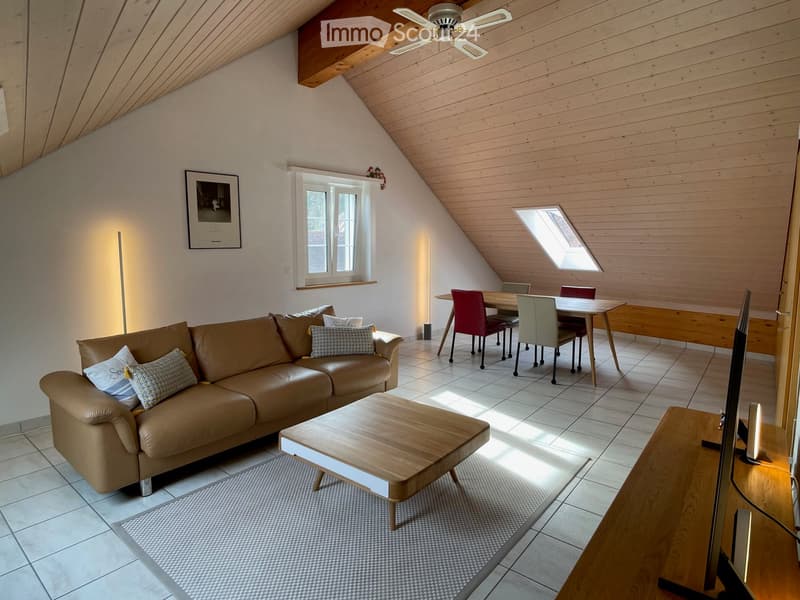 Spacious living and dining area with a brown leather sofa, wooden coffee table, dining table with chairs, and a television. The room features wooden beams on the ceiling and large windows allowing natural light to fill the space.
