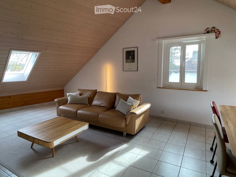 Modern living room with a brown leather sofa, wooden coffee table, and dining area with chairs and table. The room features wooden slanted ceilings, large windows, and a skylight.