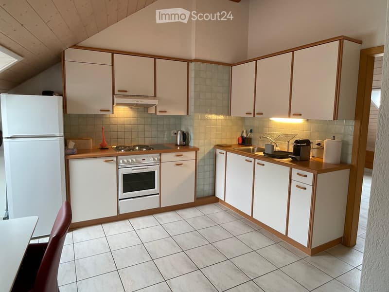 Modern kitchen with white cabinets, wooden countertops, and stainless steel appliances including a refrigerator, oven, and coffee maker. The space features tiled flooring and a small dining area with a chair.