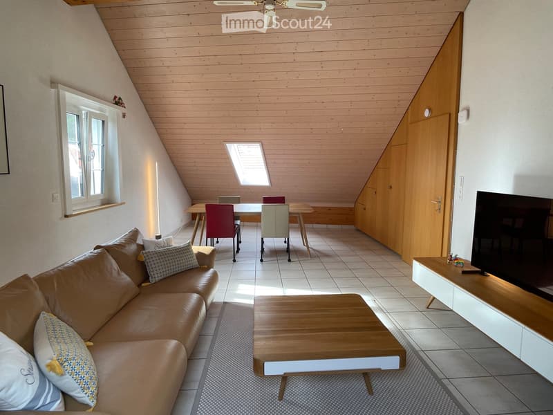 Bright living room with a slanted wooden ceiling, large windows, and skylight. Features a brown leather sofa, wooden coffee table, dining area with chairs, and a TV on a wooden stand.