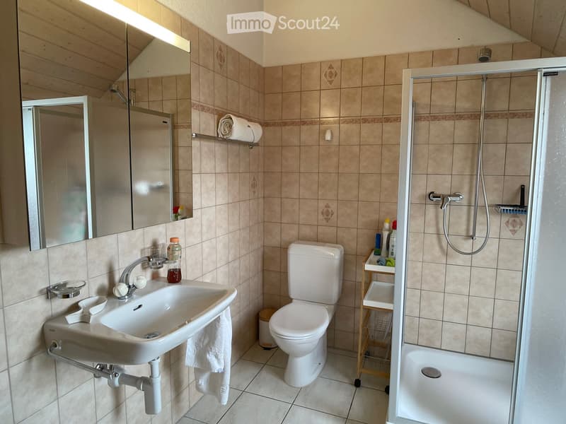 Bathroom with beige tiled walls and floor, white sink, toilet, mirror, shower, and a small cart with cleaning supplies.