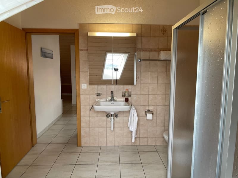 Bathroom with tiled walls and floor, a white sink with a mirror above it, a towel hanging on the wall, a toilet, and a shower area with a glass door.