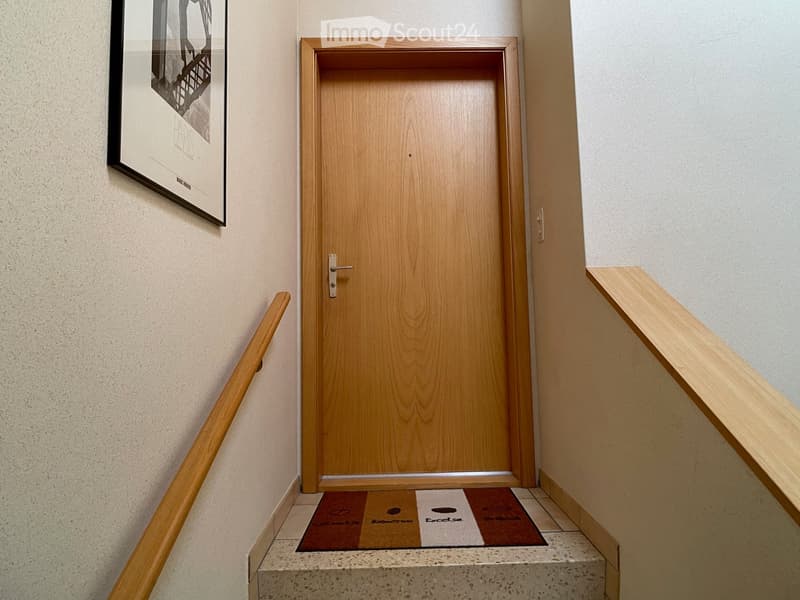 Wooden door with a silver handle, flanked by wooden handrails on both sides, with a small mat featuring a logo in front of the door.