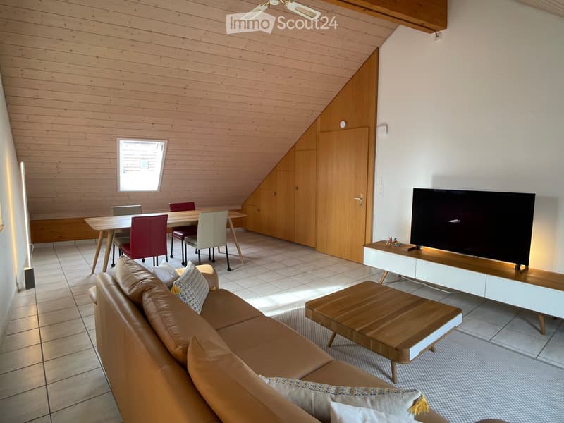 Modern living room with a slanted wooden ceiling, tiled floor, and large window. Features a brown leather sofa, wooden coffee table, flat-screen TV on a white stand, and a dining area with a table and chairs.