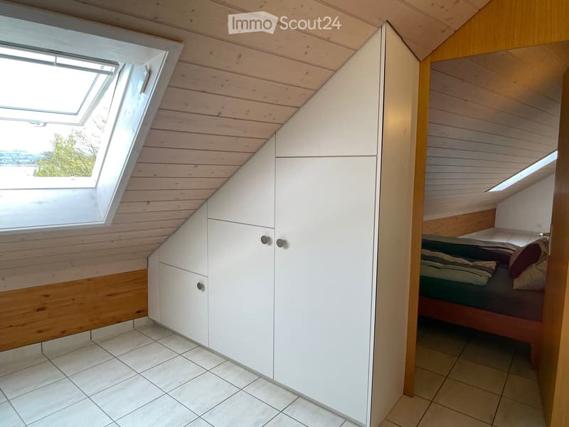 Cozy attic bedroom with wooden walls and ceiling, large skylight window, built-in white wardrobe, and a neatly made bed.