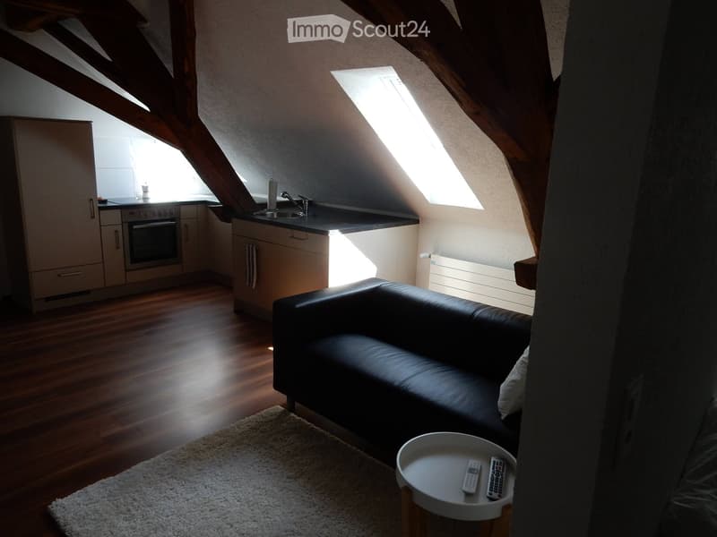 A modern, open-concept attic space with exposed wooden beams, a black leather sofa, a small coffee table, and a compact kitchen area with cabinets, an oven, and a sink.