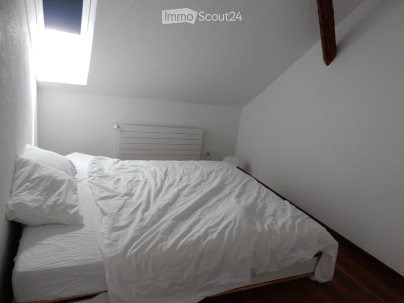 A small, cozy bedroom with a single bed, white bedding, and a radiator against the wall. The room has slanted ceilings and a small window letting in natural light.